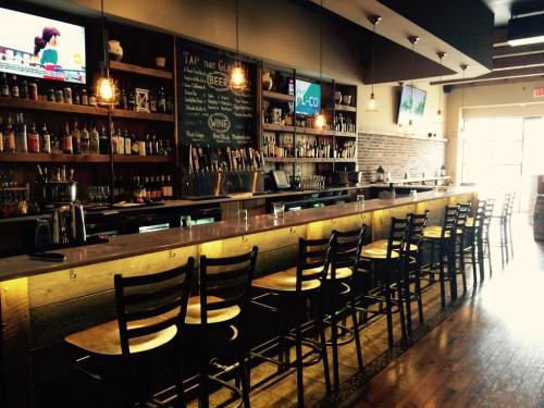 A modern bar interior with a long wooden counter, high stools, shelves of drinks, and TVs displaying sports.