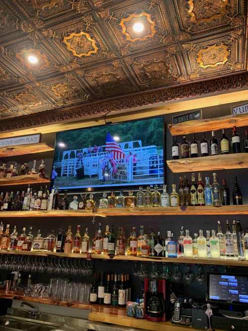 A bar with a wooden shelf displaying various bottles, featuring a TV showing a rodeo scene with an American flag.