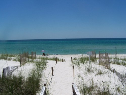 A sandy path leads to a tranquil beach with turquoise waters and a clear blue sky, framed by grass and wooden fences.
