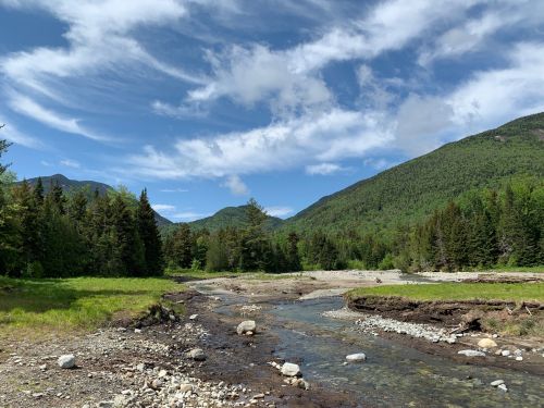 A serene landscape featuring a winding stream, lush green hills, and a blue sky with scattered clouds.