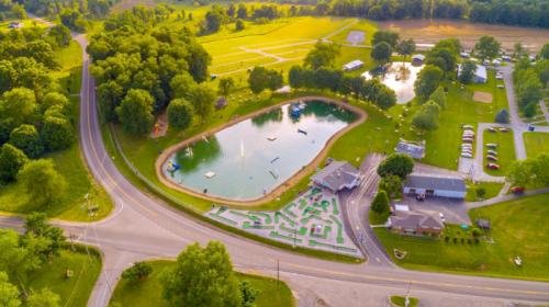 Aerial view of a park with a pond, green fields, and recreational facilities, surrounded by trees and a road.