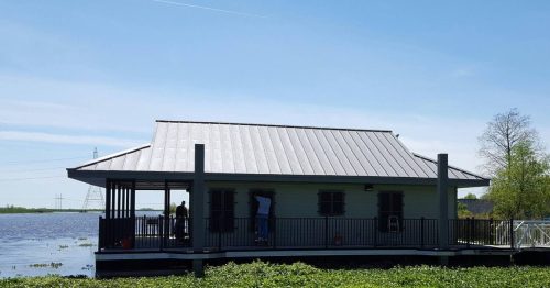 A small, modern house with a metal roof sits on stilts by a calm body of water, surrounded by greenery under a clear blue sky.