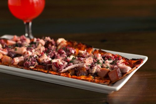 A rectangular flatbread topped with chopped meats and herbs, served alongside a glass of red beverage.