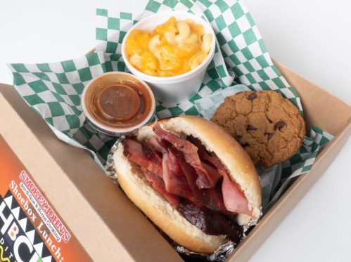 A takeout box with a sandwich filled with meats, macaroni and cheese, a cookie, and a small cup of sauce.