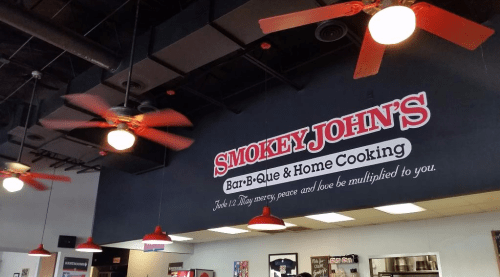 Interior of Smokey John's Bar-B-Que, featuring ceiling fans and a prominent sign with the restaurant's name and slogan.