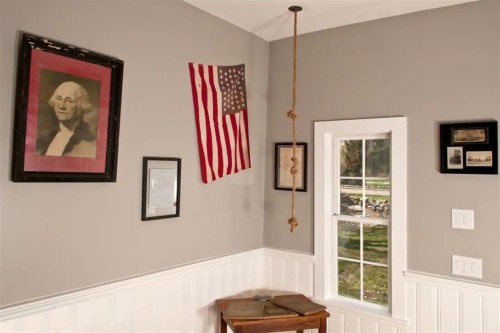 A corner of a room featuring a portrait, an American flag, and framed pictures on gray walls, with a window and table.