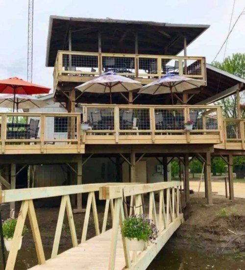 A multi-level wooden structure with a deck, umbrellas, and a ramp leading to it, surrounded by greenery.