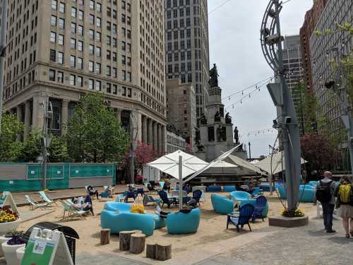 A city square with sandy areas, blue seating, and people relaxing, surrounded by tall buildings and greenery.