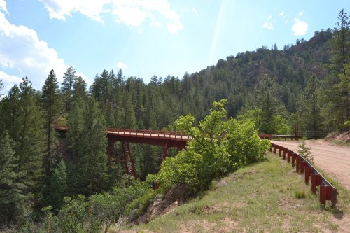 A red bridge spans a forested valley, surrounded by tall trees and a dirt road under a bright blue sky.