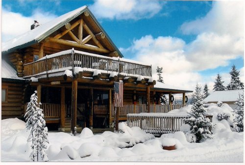 A cozy log cabin covered in snow, with a balcony and American flag, surrounded by snow-laden trees under a blue sky.