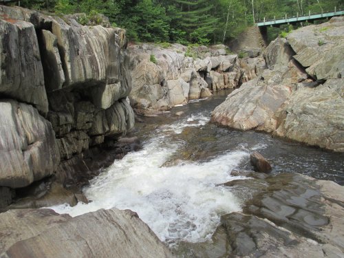 A rocky riverbed with flowing water surrounded by lush greenery and a bridge in the background.