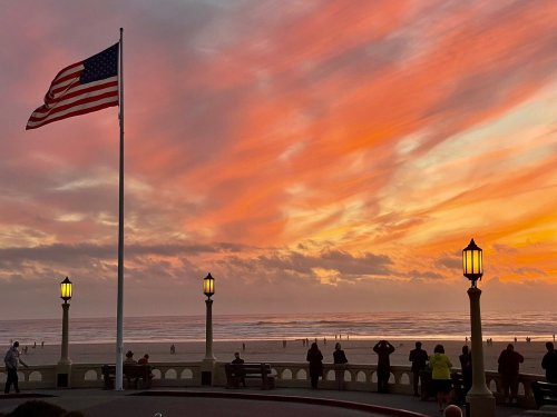 A vibrant sunset over the ocean, with people watching and an American flag waving in the foreground.