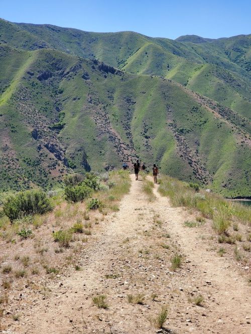 Two hikers walking along a dirt path with lush green mountains in the background under a clear blue sky.