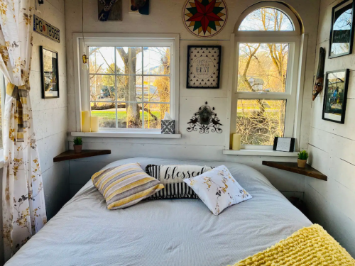 Cozy bedroom with a bed, decorative pillows, and large windows letting in natural light. Walls adorned with framed art.
