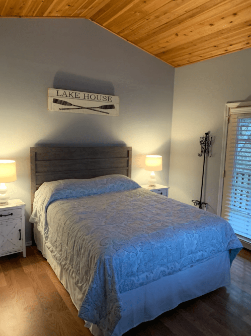 Cozy bedroom with a wooden ceiling, a queen bed, nightstands, and a "Lake House" sign on the wall.