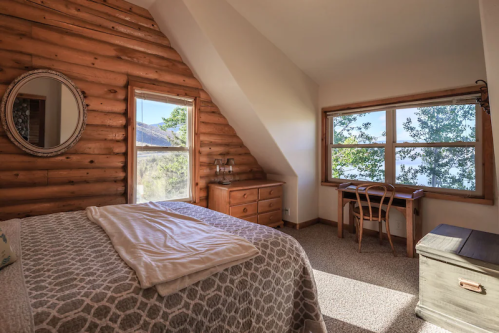 Cozy bedroom with wooden walls, a large window, a bed with a blanket, a dresser, and a small desk by the window.