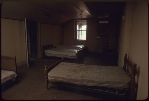 A dimly lit room with three unmade beds and a window, featuring plain walls and wooden flooring.