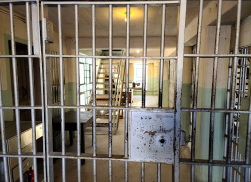 View through a barred prison cell door, revealing a dimly lit interior with stairs and empty furniture.