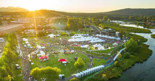 Aerial view of a lively outdoor festival with tents, crowds, and a sunset over a scenic landscape.