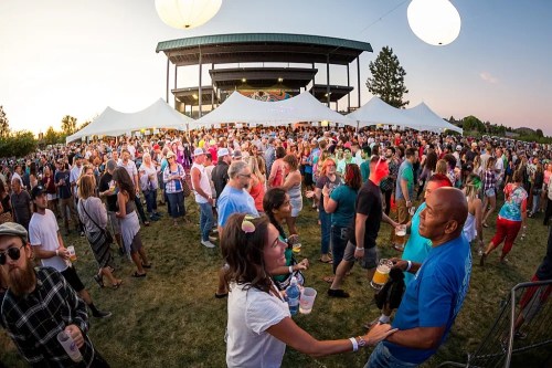 A lively outdoor festival scene with a large crowd enjoying drinks, music, and colorful decorations at sunset.