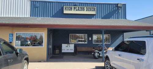 Exterior of High Plains Diner with a sign, parking area, and a motorcycle in front. Clear blue sky above.