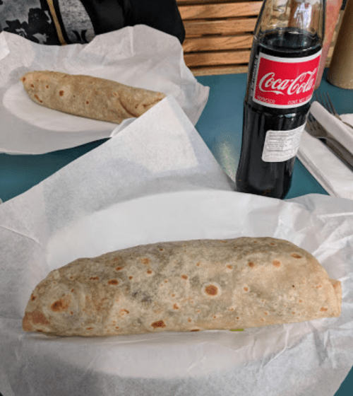 Two burritos on white paper plates, with a bottle of Coca-Cola in the background.