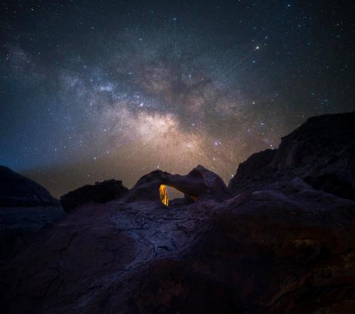 A stunning night sky filled with stars and the Milky Way, framed by a rocky arch in a dark landscape.