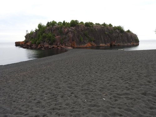 A small rocky island surrounded by calm water, with a sandy beach in the foreground and greenery on the island.