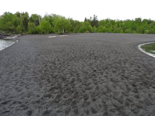 A sandy beach with dark gray sand, surrounded by lush green trees and a calm water edge.