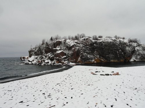 A snowy beach scene with a rocky cliff and calm water under a cloudy sky.