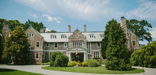 A large, elegant stone mansion surrounded by lush greenery and trees under a clear blue sky.