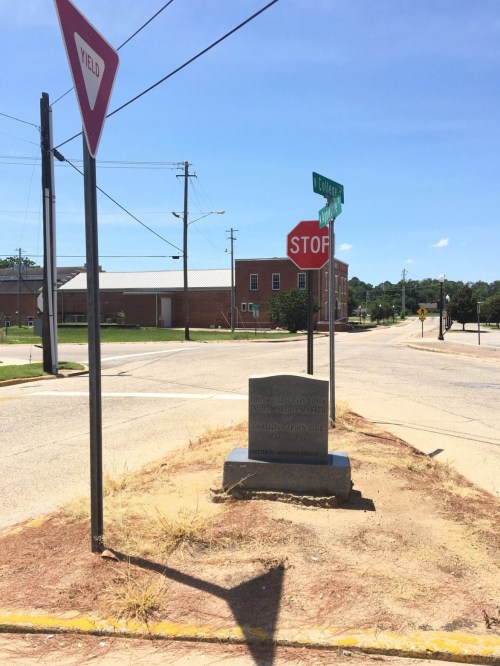 A street corner with a stop sign, yield sign, and a stone monument on a grassy area. Buildings in the background.