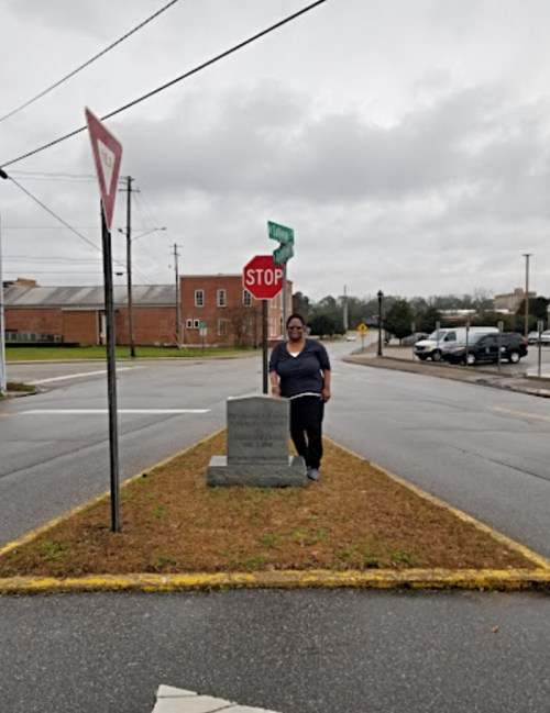 A person stands next to a stop sign and a monument on a grassy median at an intersection on a cloudy day.