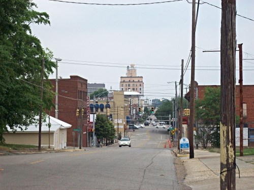A view of a quiet street lined with buildings, leading to a distant city skyline under a cloudy sky.