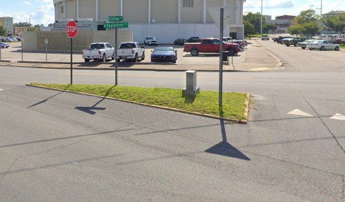 A street corner with a stop sign, grassy median, and a parking lot filled with cars in the background.
