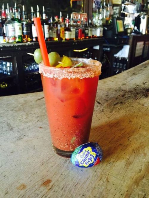 A glass of Bloody Mary with a salted rim, garnished with olives, next to a Cadbury Crème Egg on a wooden bar.