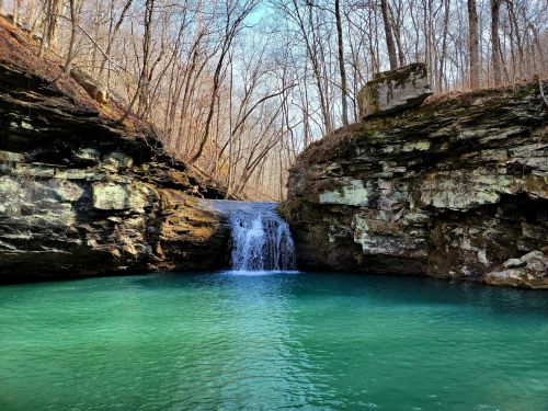 A serene waterfall cascades into a turquoise pool, surrounded by rocky cliffs and bare trees in a tranquil forest setting.