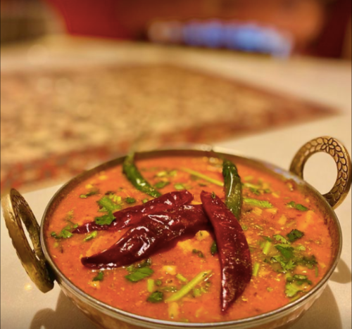 A bowl of vibrant red curry garnished with green chilies and dried red chilies, set on a decorative table.