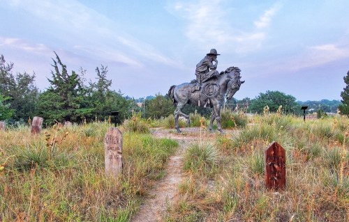 A bronze statue of a cowboy on horseback surrounded by tall grass and wooden markers in a serene landscape.