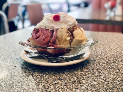 A glass dish of ice cream with three scoops, topped with whipped cream and a cherry, on a speckled table.
