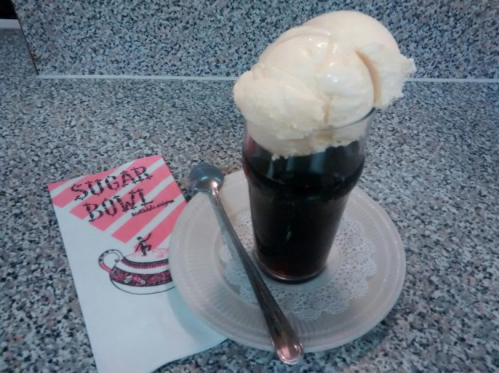 A glass of root beer float topped with vanilla ice cream, served on a plate with a spoon and a menu beside it.