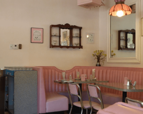 A retro diner interior with pink booths, a mirrored wall, and a vintage light fixture, creating a cozy atmosphere.