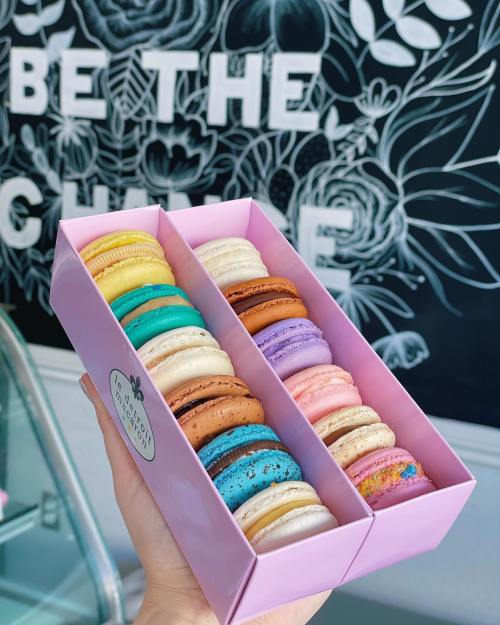 A hand holding a pink box filled with colorful macarons against a decorative wall.