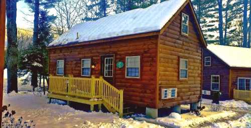 A cozy wooden cabin with a yellow porch, surrounded by snow and trees, under a clear sky.