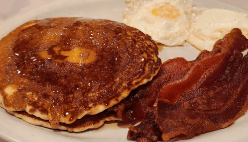 A plate with two pancakes drizzled with syrup, two fried eggs, and crispy bacon strips.