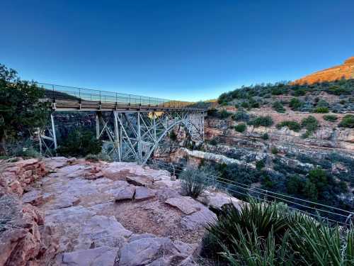 A metal bridge spans a rocky canyon, surrounded by greenery and red rock formations under a clear blue sky.