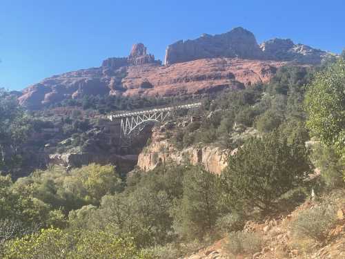 A scenic view of red rock formations and a bridge nestled among trees under a clear blue sky.