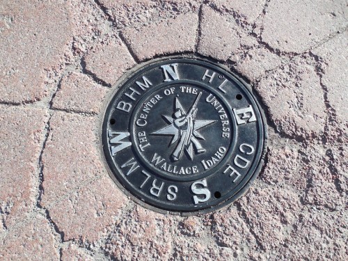 A metal marker on the ground labeled "The Center of the Universe" in Wallace, Idaho, with directional letters and a star design.