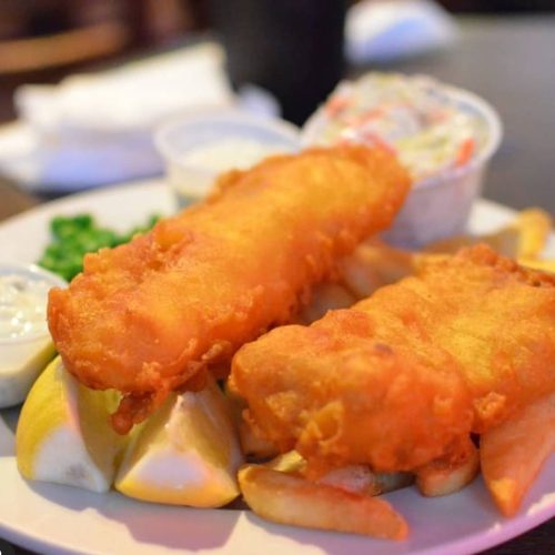 A plate of golden fried fish fillets with lemon wedges, fries, and coleslaw on the side.