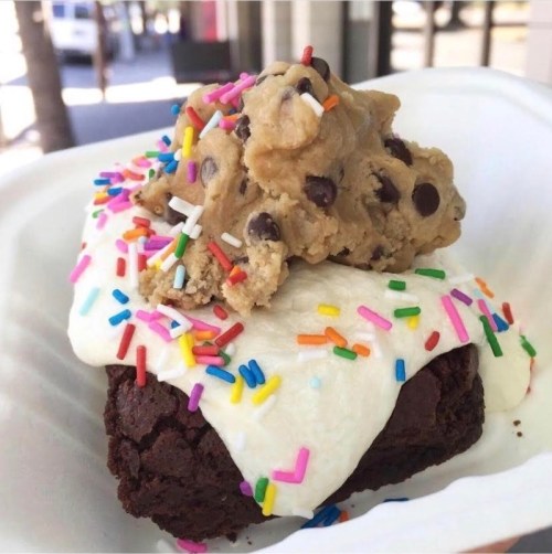 A decadent brownie topped with cream, cookie dough, and colorful sprinkles on a white plate.